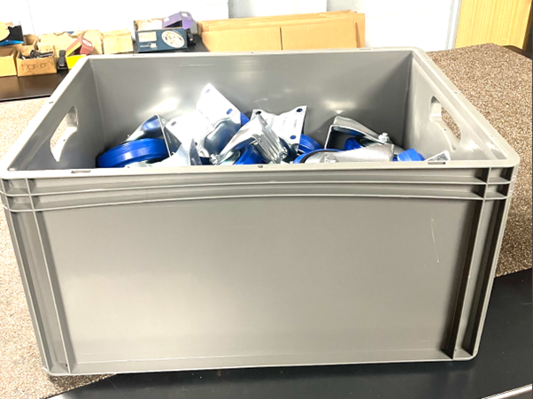 Grey plastic tote storage bin filled with blue industrial castor components placed on warehouse workbench for organised parts handling.