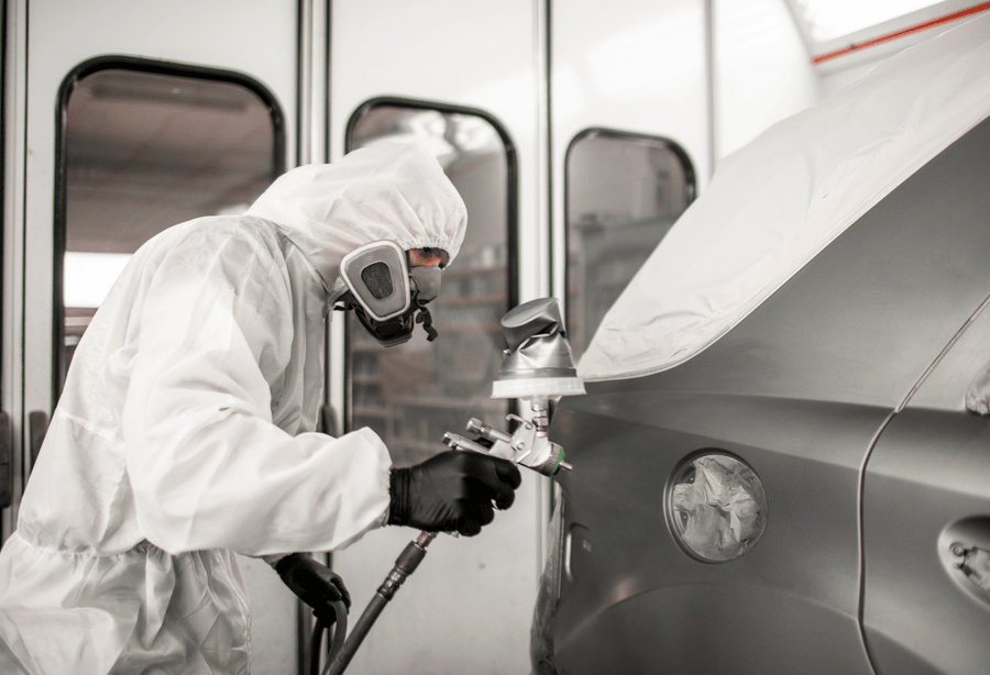 Person in full protective gear using a spray gun to coat a car inside an automotive paint booth.