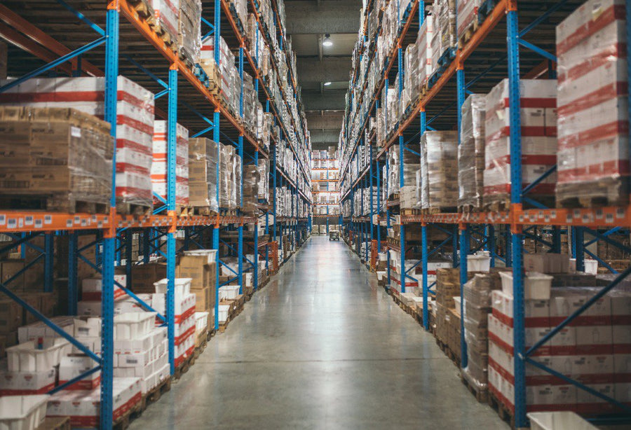 Interior of a large warehouse with tall shelves stacked with boxes and pallets, showing a wide aisle used for transporting goods.
