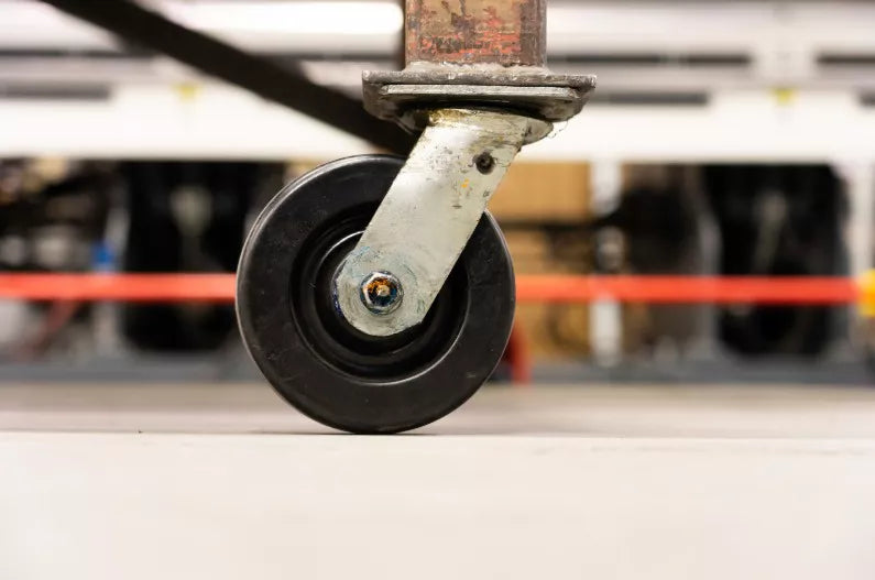 Close-up of a black trolley castor wheel mounted on a metal frame, set in a blurred industrial workshop environment.