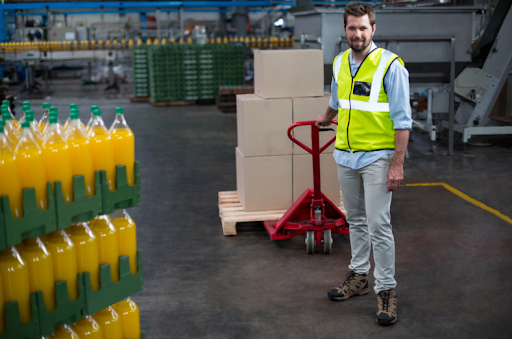 Warehouse worker in a high-visibility vest standing next to a manual pallet jack and stacked boxes in a beverage distribution facility.